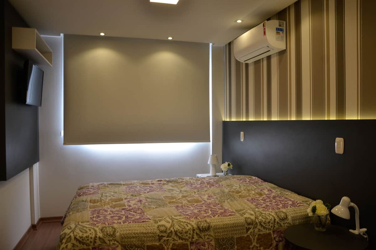 A hotel room with a bed, a TV mounted on the wall, an air conditioning unit, a lamp, and a small table with a vase.
