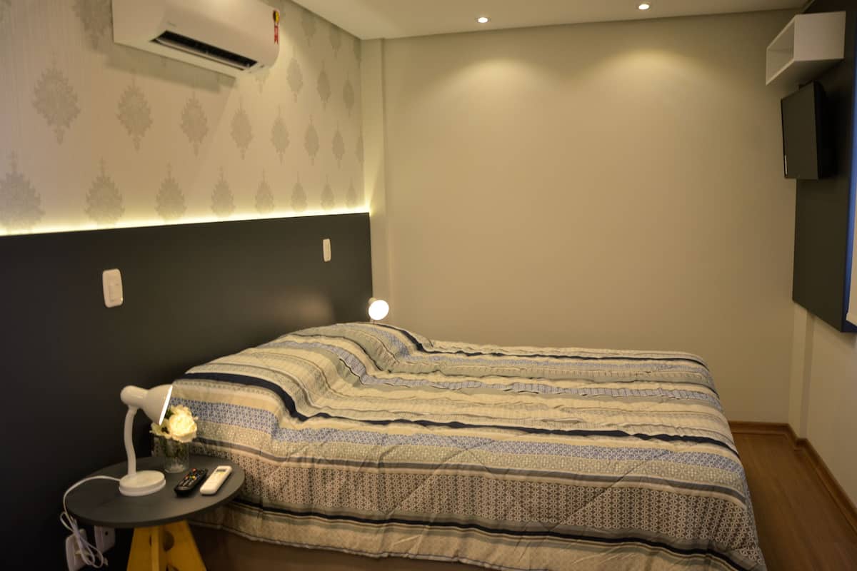 A neatly made bed with a patterned bedspread, a bedside table with a lamp and remote controls, and a wall-mounted air conditioner.