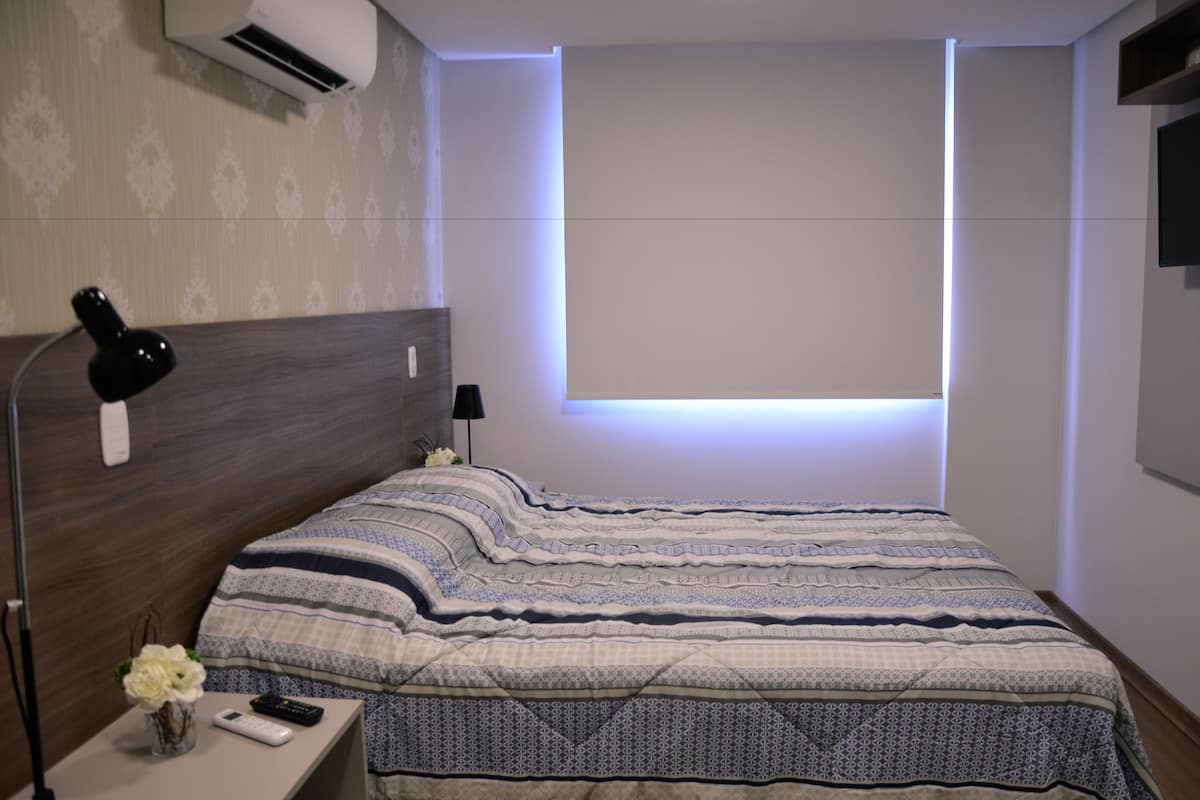 A neatly made bed with a patterned bedspread, a bedside lamp, a remote control, and a vase with flowers on a nightstand.