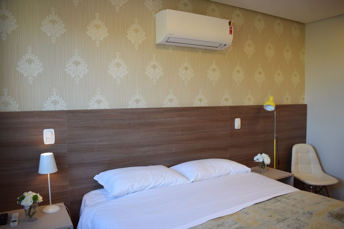 A hotel room with a wooden headboard, a bed with white bedding, a nightstand with a lamp and a vase of flowers, and an air conditioning unit on the wall.