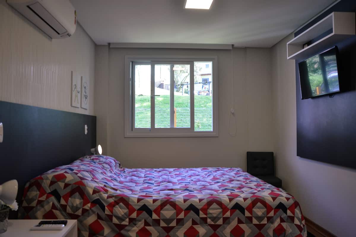A bedroom with a bed, a window, a TV, and a wall-mounted air conditioner.