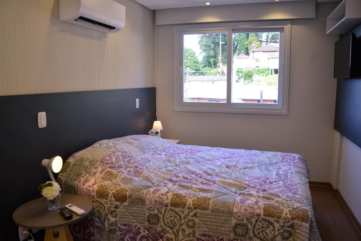 A bedroom with a large bed, a bedside table with a lamp, a small table with a vase and remote control, and a window with a view of the outside.