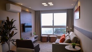Una sala moderna con sofá, sillones, una mesa de centro y un televisor montado en la pared. Hay plantas en macetas y un ventanal con cortinas.