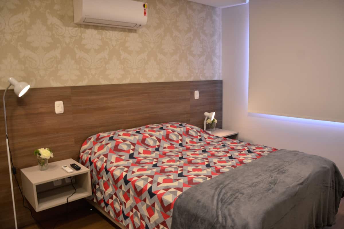 A neatly arranged hotel room with a wooden headboard, a patterned bedspread, a bedside table with a lamp, and a small vase with flowers.