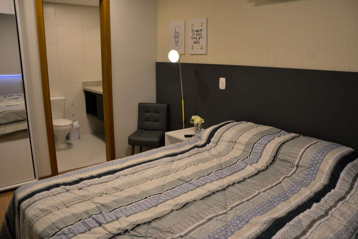 A bedroom with a bed, a nightstand, a chair, a desk, a lamp, and a wall with two framed pictures.