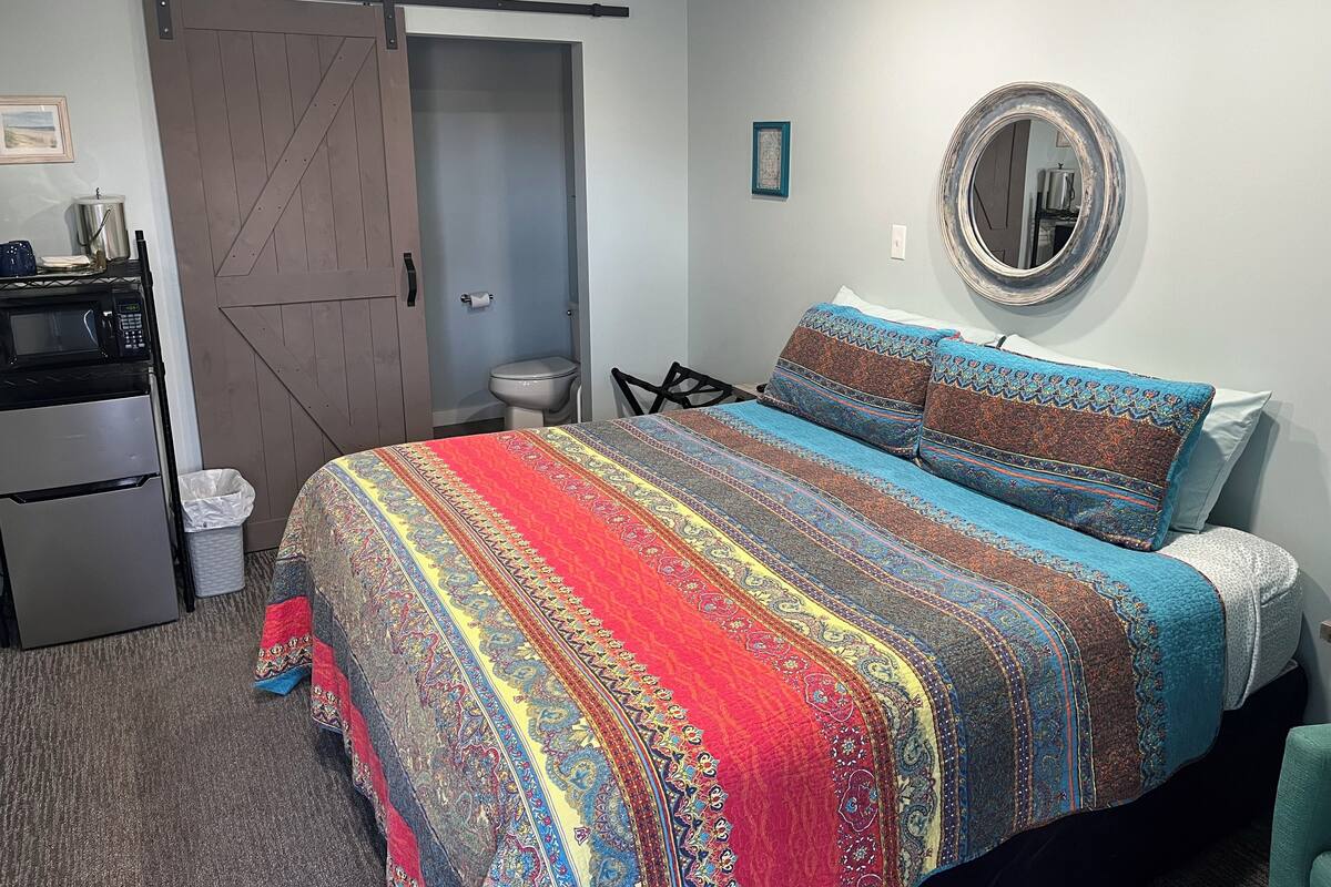 A bedroom with a bed, a dresser, a microwave, a refrigerator, a sliding door, a toilet, and a mirror on the wall.