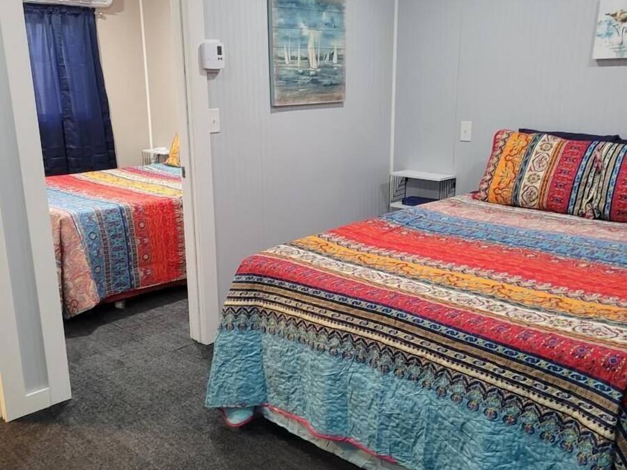 A bedroom with two beds, a small table, and a chair. There is a painting on the wall and an air conditioning unit in the corner.