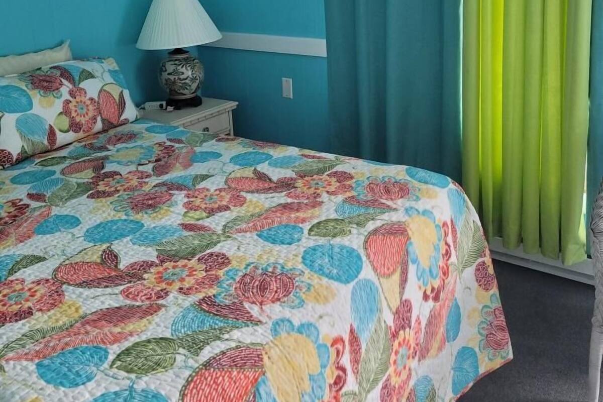 A bedroom with a floral bedspread, a nightstand with a lamp, and a window with green curtains.