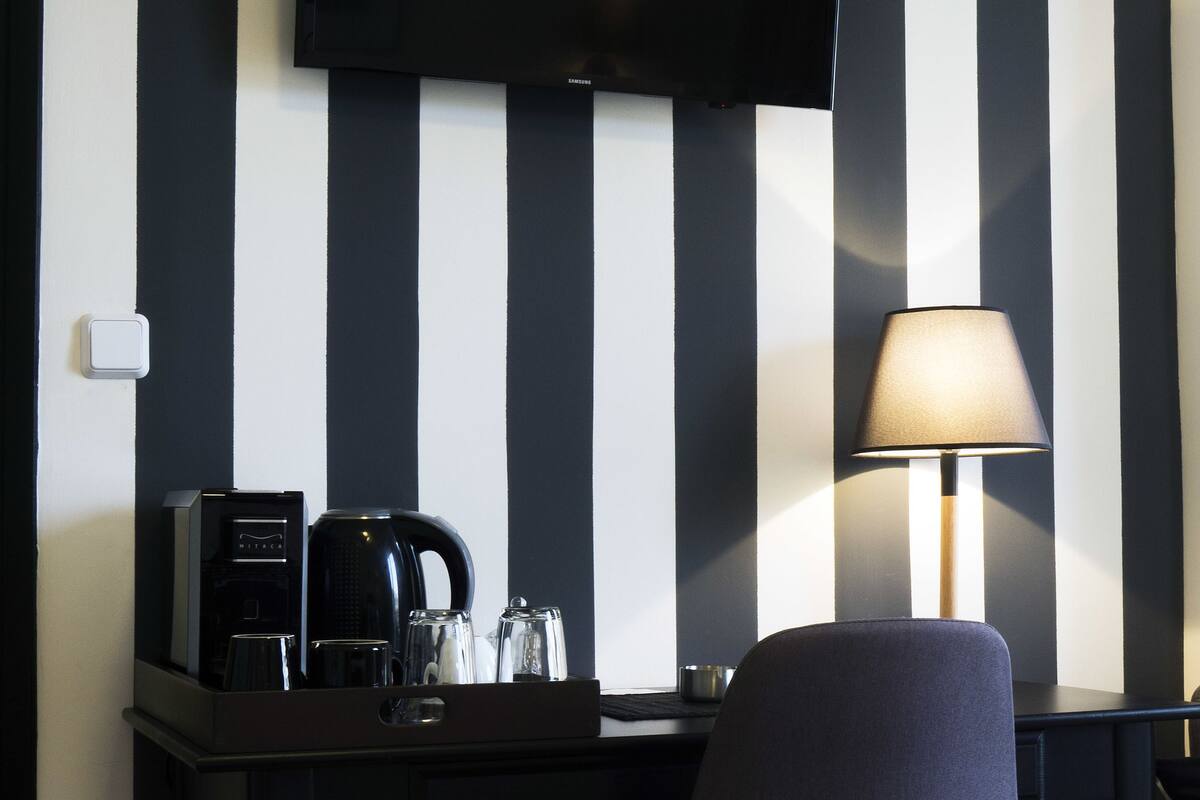 Ein Hotelzimmer mit einer schwarz-weiß gestreiften Wand, einem flachbildfernseher über einem Schreibtisch mit Kaffeemaschine und Gläsern, und einem Sessel mit Lampe.