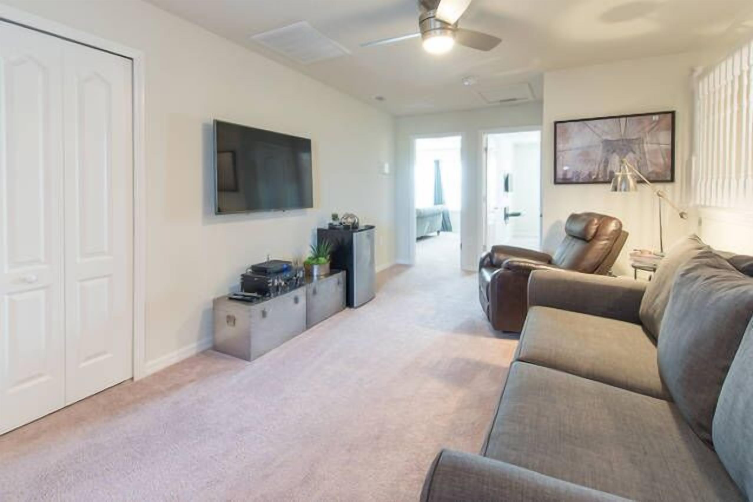 A living room with a grey sofa, a TV mounted on the wall, a ceiling fan, and a door on the left.