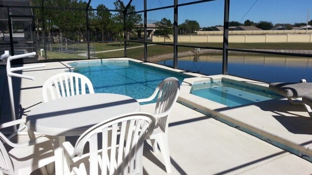An outdoor swimming pool area with white chairs and tables, surrounded by a glass enclosure. - Captain Jack's Getaway (Kissimmee)