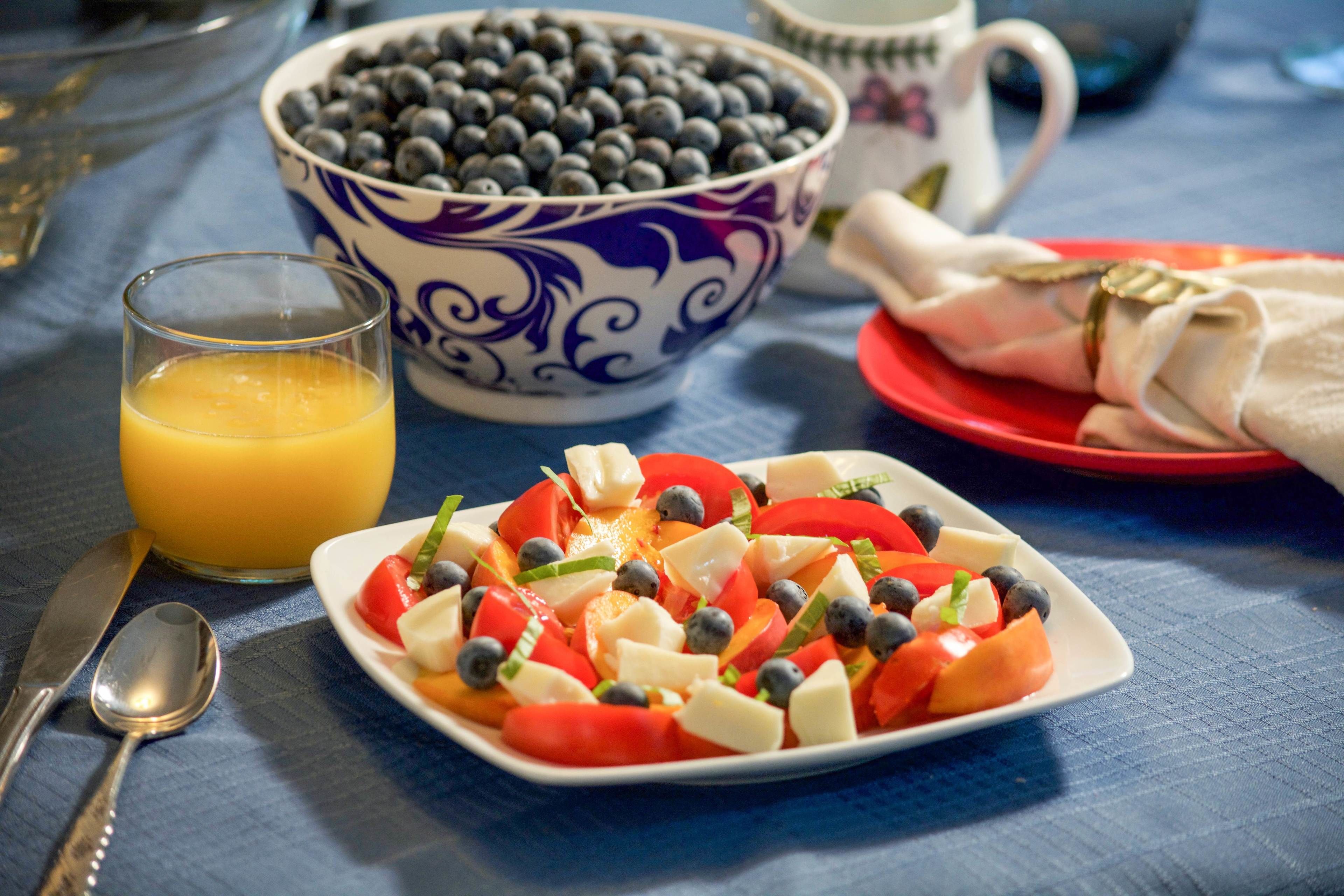 A breakfast setting with a plate of fruit salad, a bowl of blueberries, a glass of orange juice, and a coffee cup on a blue tablecloth.
