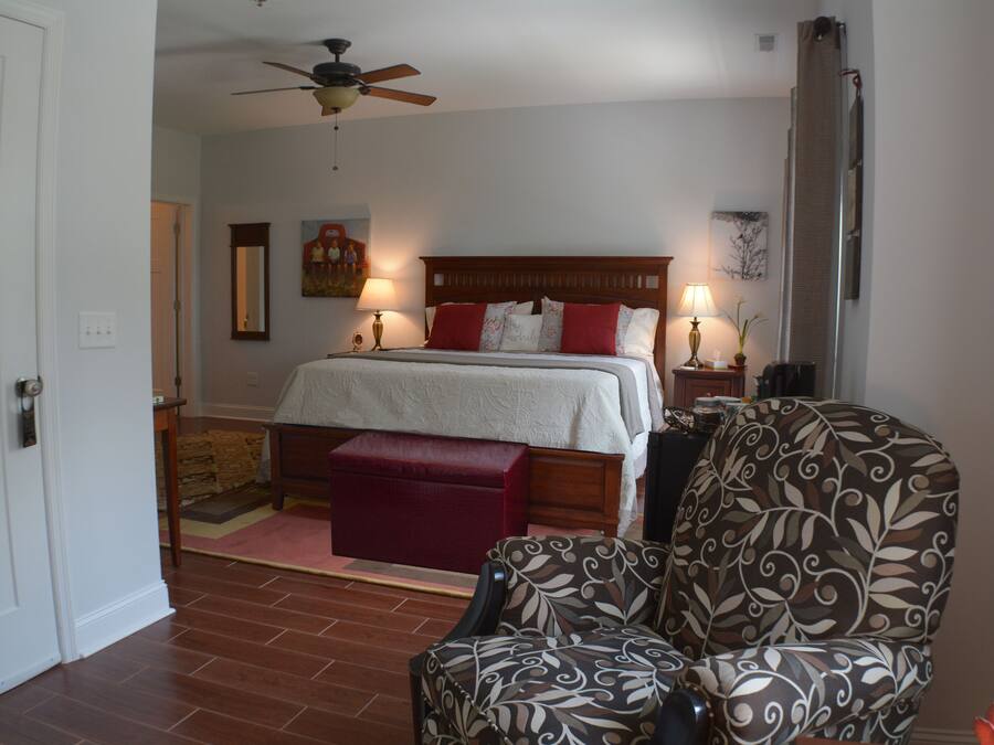 Quarto com cama, um banquinho vermelho, poltrona estampada, ventilador de teto e duas luminárias de cabeceira.