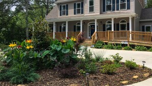 A two-story house with a porch, a front door with a wreath, and a garden with various plants and flowers. - St Francis Cottage (Chattanooga)