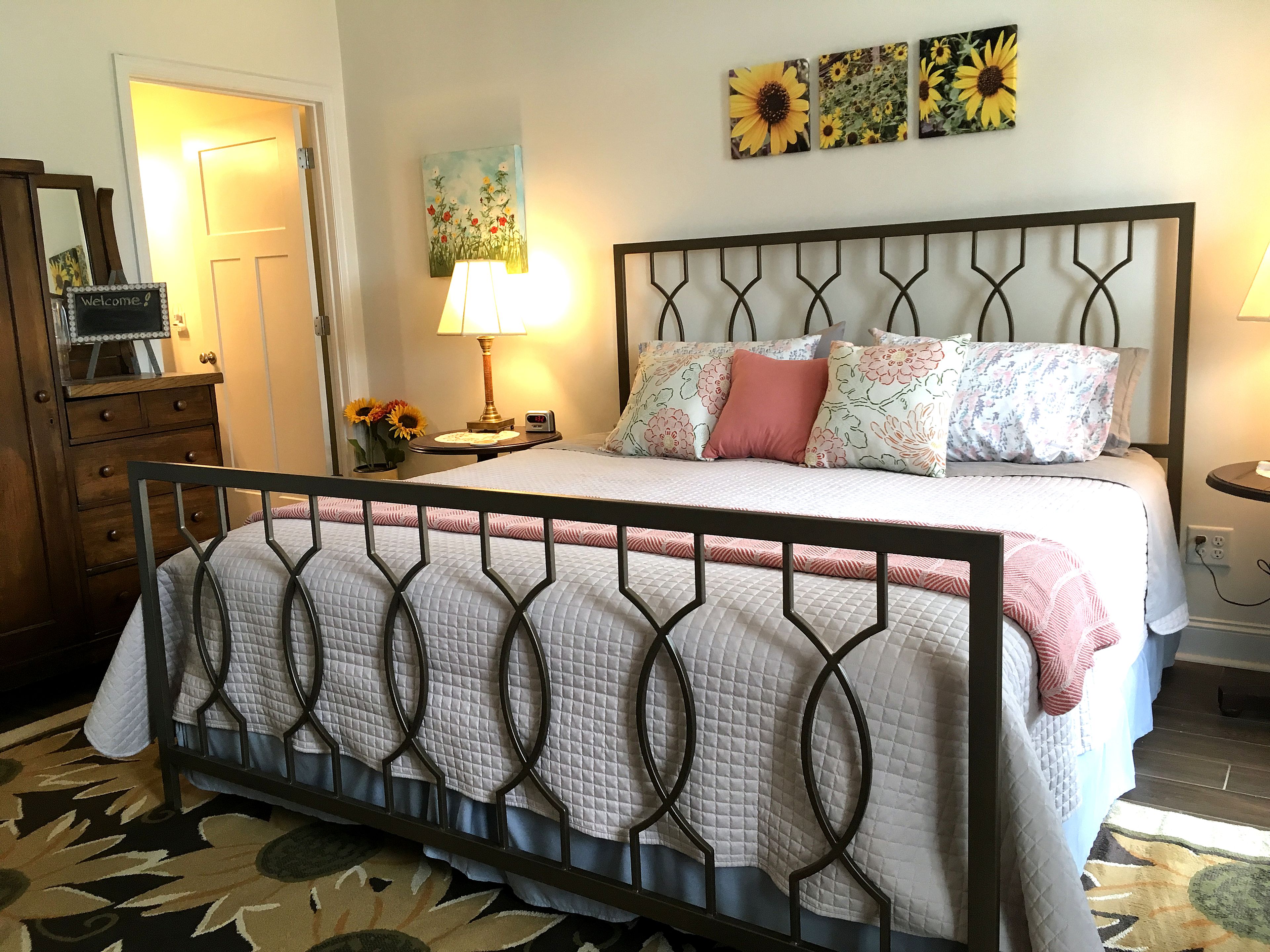Un lit bien fait, avec une tête de lit en métal, une table de chevet avec une lampe et un mur orné de trois tableaux représentant des tournesols.