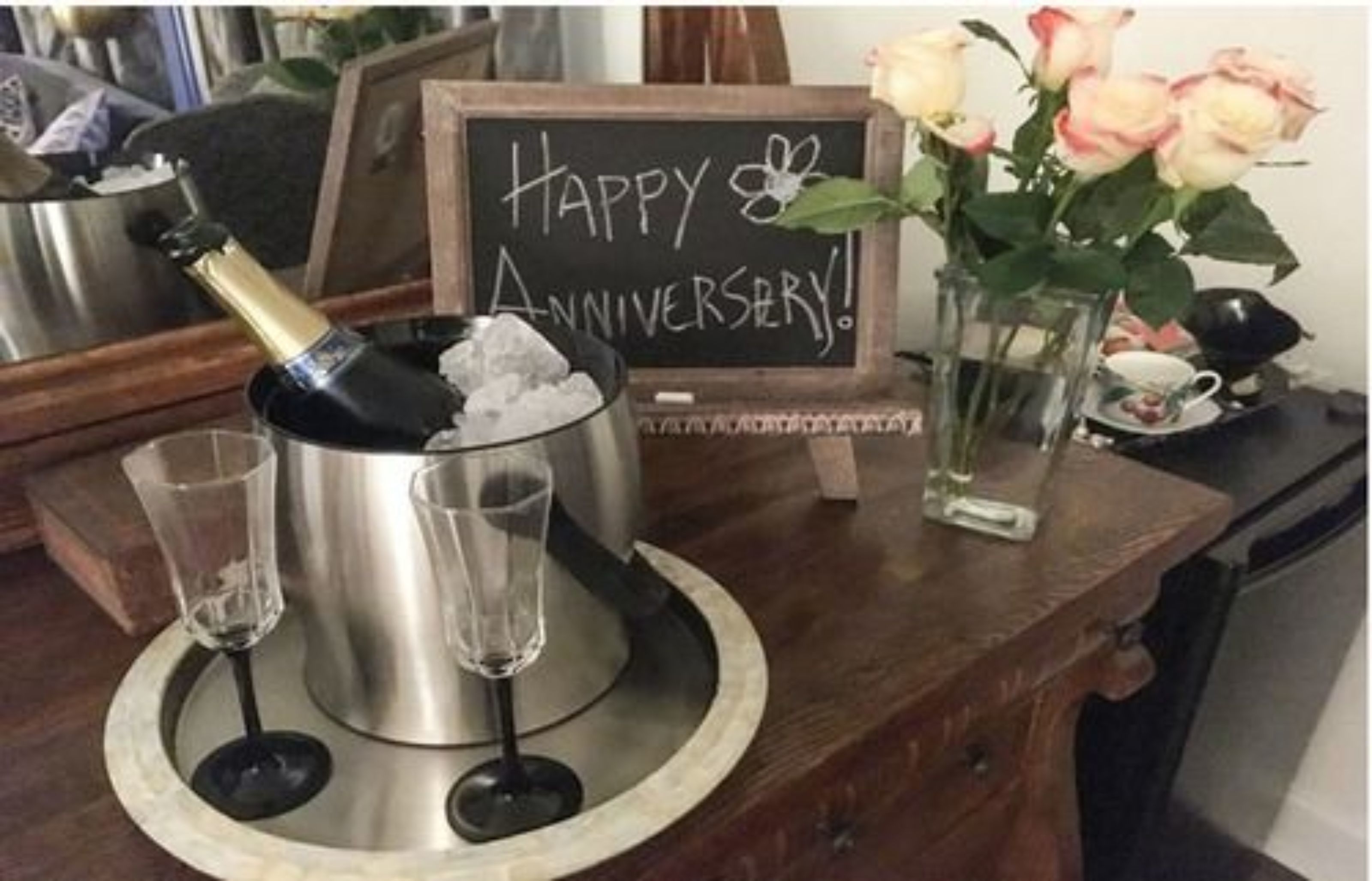 A table with a champagne bottle, two glasses, a chalkboard sign saying "Happy Anniversary!", and a vase with roses.