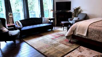 A room with a dark-colored sofa, a chair, a bed with a patterned bedspread, a flat-screen TV on a stand, and a large window with curtains.