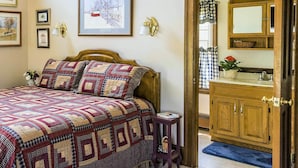 A bedroom with a wooden bed, a quilt, framed pictures on the wall, a nightstand with a vase, and a view into the bathroom with a sink and mirror. - Red Cardinal Bed and Breakfast (Carlisle)