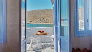 A balcony with white wicker chairs and a small round table, overlooking a coastal area with a clear blue sky and a rocky hillside in the background. - Amo Suites & Spa (Folegandros)