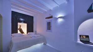 A modern bedroom with a bed, a nightstand, a laptop on a shelf, and a wall-mounted light. - Amo Suites & Spa (Folegandros)