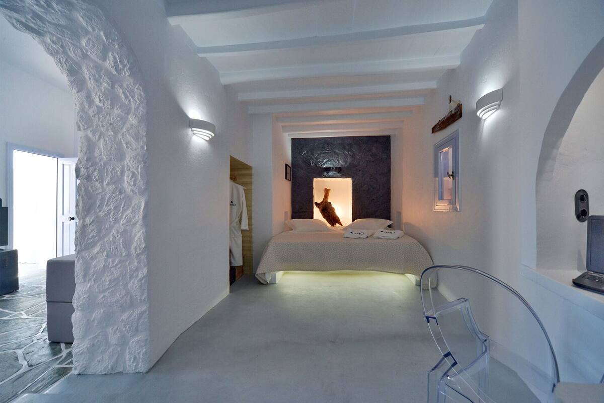A minimalist hotel room with a bed, a wall-mounted light, and a view of the sea.