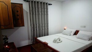 A bedroom with a single bed, a wooden nightstand, a small table, and a curtain with a floral pattern. - Villa Peacock (Hikkaduwa)