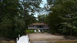 A lakeside cabin with a dock extending into the water, surrounded by dense trees. - Sandy waterfront, grass yard, 40' private dock. Foliage week special-Book now! (Moultonborough)