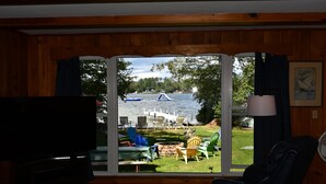 A living room with a large window overlooking a lake, outdoor seating, and a television. - Sandy waterfront, grass yard, 40' private dock. Foliage week special-Book now! (Moultonborough)