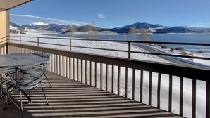 A balcony with a metal chair and a table, overlooking a snowy landscape with mountains and a lake.