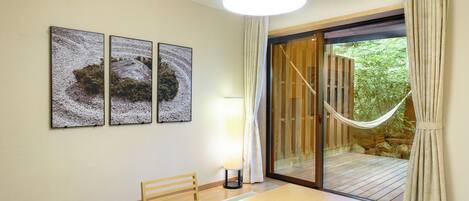 Una habitación con una mesa de madera, dos sillas, una lámpara y vistas a un jardín a través de un ventanal corredero.