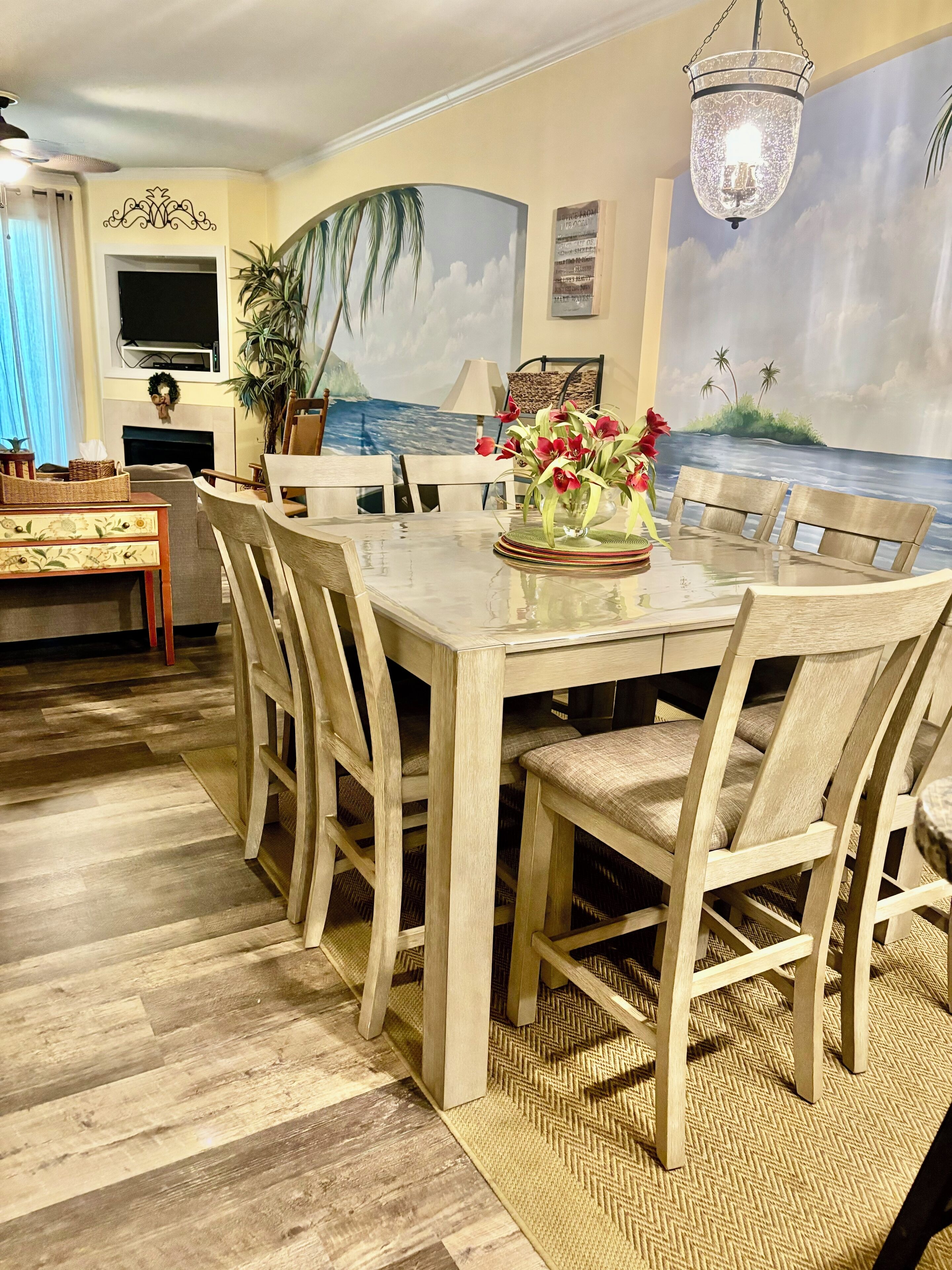 A dining area with a wooden table and chairs, a chandelier, and a mural of a beach scene on the wall.