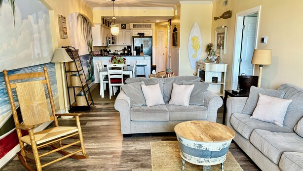 A living room with a grey sofa, a wooden rocking chair, and a coffee table. There is a kitchen in the background with white cabinets and a dining table. - ADAGIO#207 BEAUTIFUL 3 BED/3 BATH OCEAN BLOCK CONDO W/INDOOR POOL STEPS TO BEACH (Ocean City)