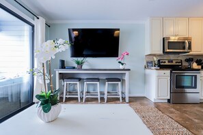 A modern kitchen with a flat-screen TV, white cabinets, a microwave, and a stainless steel oven. There are three bar stools and a potted plant with white orchids.