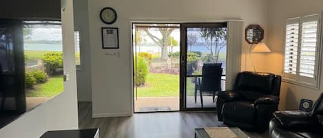 A living room with a black leather sofa, a coffee table, and a TV. There is a sliding glass door leading to a patio with a view of the outdoors.