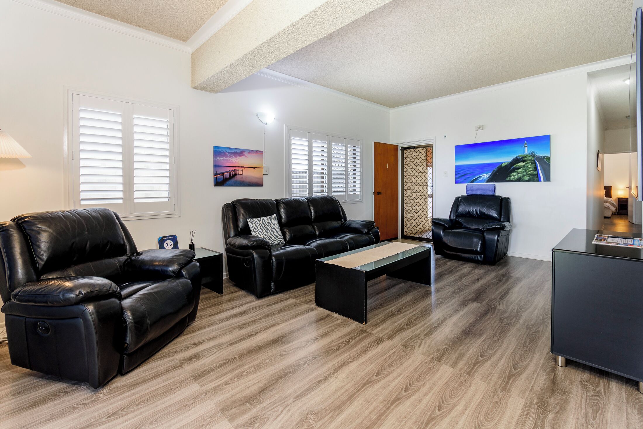 A modern living room with a leather sofa, a recliner, and a coffee table. There are two windows with blinds, a painting on the wall, and a door with a mesh screen.