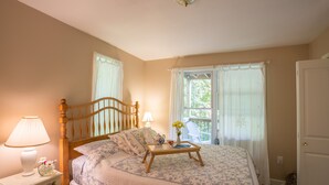 A bedroom with a wooden bed, a bedside table with a lamp, a small table with flowers, and a window with curtains. -  Resort Mountain Home with  Private Hot tub Walk to Pools, Pond WIFI (Campton)