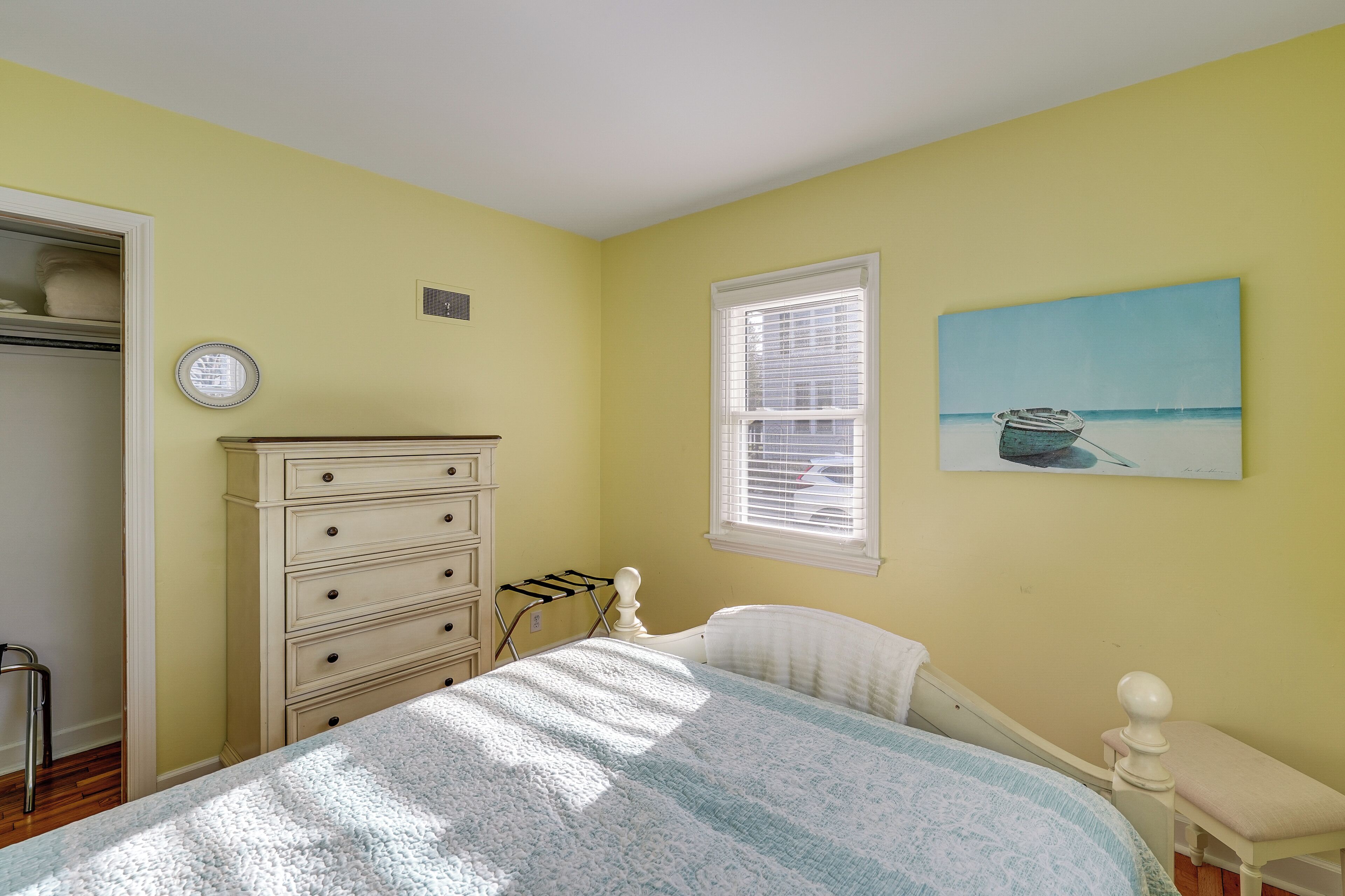 Une chambre à coucher comprenant un lit, une commode, une fenêtre avec des stores, un tableau au mur et une armoire avec une porte.