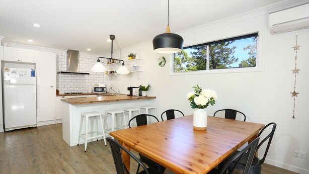 Una cocina moderna con armarios blancos, una mesa de comedor de madera y sillas negras. Se ve una ventana con vista a árboles y una decoración con forma de estrella colgante.