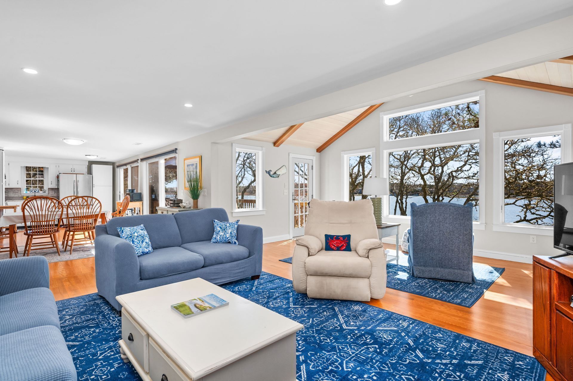 Un amplio salón con un sofá azul, un sillón beige y otro sillón azul. Hay una mesa de centro blanca con una revista encima. La habitación tiene ventanas grandes con vistas a árboles y una masa de agua.