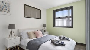 Een slaapkamer met een bed, twee nachtkastjes, een raam met zonwering en een kunstwerk in een lijst aan de muur.