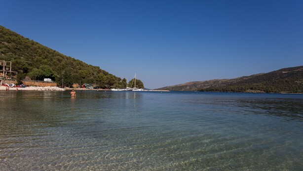 A clear lake with boats, a sandy beach, and a forested hill.