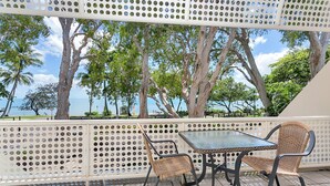 Une terrasse extérieure avec une table et des chaises, entourée d’une clôture blanche ajourée, offrant une vue panoramique sur des arbres et une étendue d’eau.
