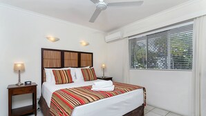 Une chambre d’hôtel avec un grand lit, deux tables de chevet avec des lampes, un ventilateur de plafond et une fenêtre avec des stores.