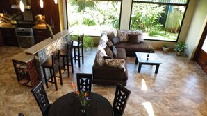 A spacious living area with a dining table, sofa, and chairs. There is a kitchen area with a stove and cabinets. Large windows provide natural light and a view of greenery outside. - Luxurious Castle, Infinity Pool And Jaw Dropping Views!  (Atenas)