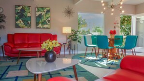 A modern living room with a red sofa, teal chairs, and a coffee table. There are two wall-mounted paintings, a floor lamp, and a large window with a view of greenery. - PS-ID #76 Stylish Mid-Century Modern Oasis with Surrounding Mountain Views (Palm Springs)