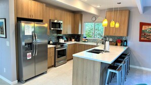 A modern kitchen with stainless steel appliances, wooden cabinets, a central island with bar stools, and pendant lighting. - PS-ID #76 Stylish Mid-Century Modern Oasis with Surrounding Mountain Views (Palm Springs)