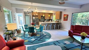 A modern living room with a red sofa, a glass dining table, and teal chairs. There is a kitchen in the background with wooden cabinets and a bar area. - PS-ID #76 Stylish Mid-Century Modern Oasis with Surrounding Mountain Views (Palm Springs)