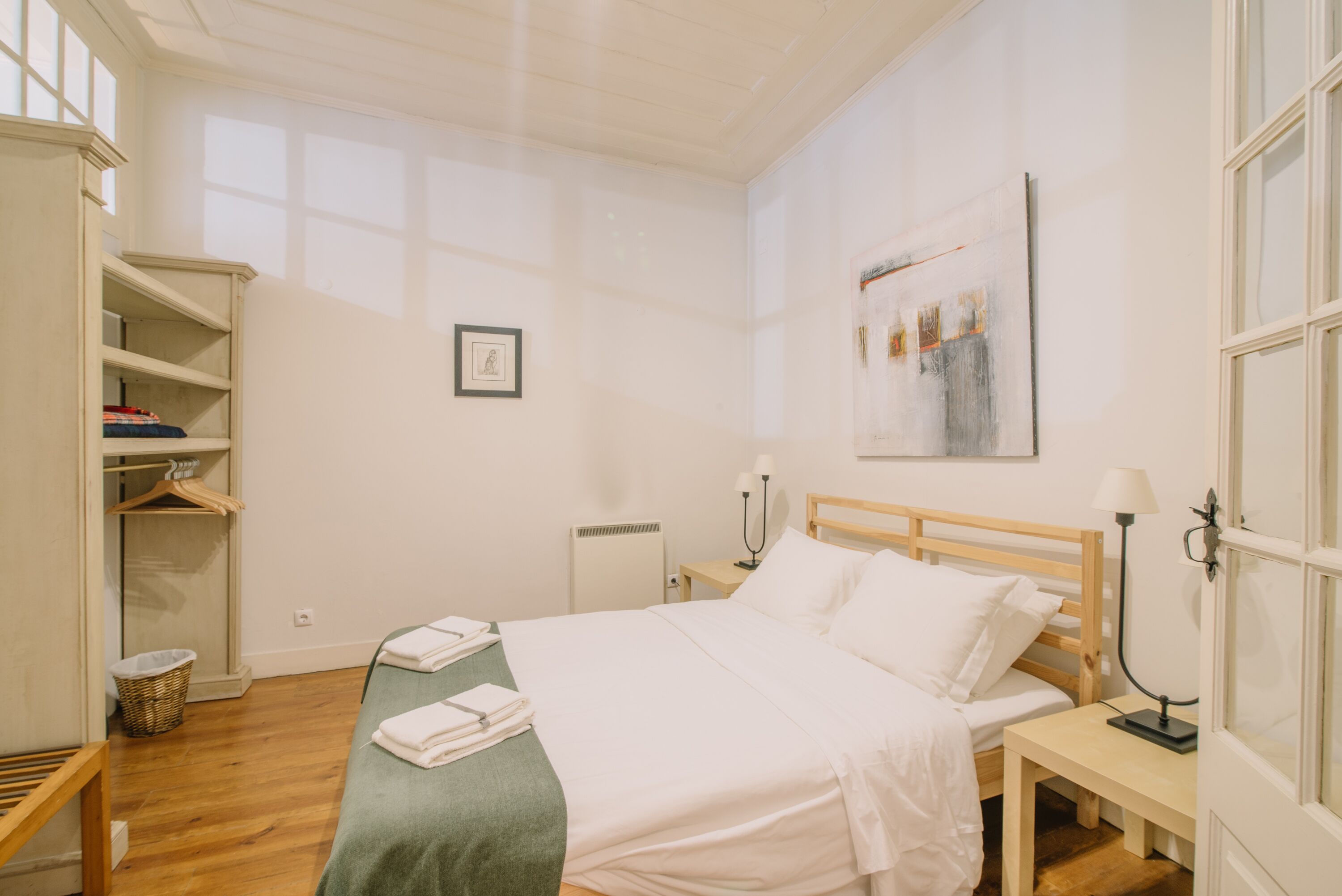 Un dormitorio ordenado con cama, mesitas de noche, lámpara, un cuadro enmarcado en la pared y una ventana.