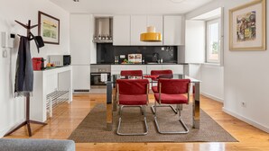 A modern kitchen with white cabinets, a black countertop, and a yellow pendant light. There is a dining area with a glass table and red chairs. - Chiado | STYLISH & GREAT VIEW | Family-Friendly (Lisboa)