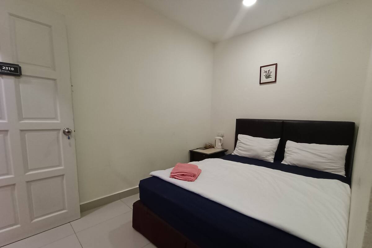 Quarto com cama, mesa de cabeceira, quadro na parede e porta com placa de número.